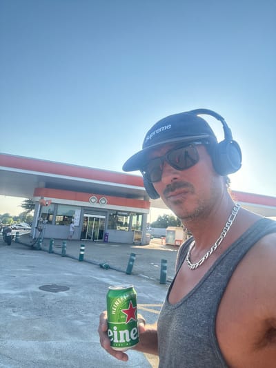 The image depicts a man standing at a gas station in Llinars del Vallès, Spain. He is wearing a grey tank top, a black baseball cap with "Supreme" printed on it, sunglasses, and headphones. He is holding a can of Heineken beer in his right hand. In the background, the gas station building, pumps, and a few cars are visible. The sky is clear and blue. The lighting suggests it is a sunny day.
