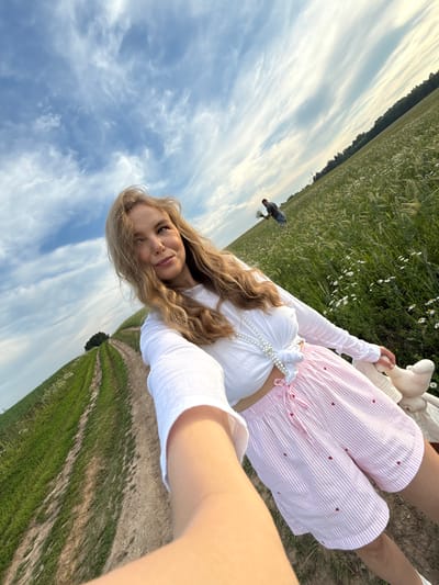 An outdoor, low-angle, selfie-style shot captures a young woman in a field. She has blonde hair, is wearing a white cropped top and pink striped shorts with red hearts, and is holding a stuffed animal. To her right is a field of green grass with white flowers and trees in the background. A dirt road with green grassy edges is to her left. In the distance, a man is walking through the field, carrying a bouquet of flowers. The sky above is blue with wispy white clouds. The location is specified as Сновицы, Russia.