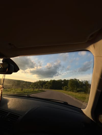 The image captures a first-person perspective from inside a vehicle, looking forward through the windshield at a rural or semi-rural landscape in Hrabovets, Ukraine, during late afternoon or early evening. The sky displays a mix of light blue areas and scattered cumulus clouds, with some clouds illuminated by warm, golden sunlight from the left, indicating the sun's low position. The foreground inside the car shows a dark dashboard, textured and partially illuminated by the same warm light on the left side, revealing air vents. A rearview mirror is visible on the upper left, from which a decorative item hangs. The beige headliner and the right A-pillar of the car are also visible, framing the upper and right portions of the view.

Outside the vehicle, an asphalt road curves gently to the right, bordered by green grass and lush trees on both sides. In the distance to the left, rolling hills covered with vegetation are visible, bathed in the soft, warm light of the setting sun. Several faint wooden posts or markers stand along the right shoulder of the road. Utility poles and wires are subtly discernible above the treeline on the right. No people or other active subjects are present; the scene depicts a tranquil natural environment observed from a stationary or moving vehicle.