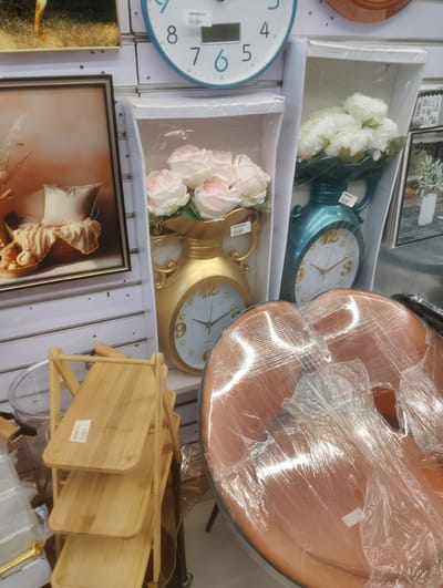 The image shows a retail display of home decor items. The subjects include decorative clocks shaped like ornate vases, one gold and the other teal. The gold clock is adorned with a bouquet of light pink and white artificial roses. The teal clock has white flowers. Both clocks have white faces with gold numerals and hands. In the background, a framed artwork depicts a soft rendering of cushions and drapery. To the left of the clocks, a wooden multi-tiered shelf unit is visible, with each shelf holding a small white label. In the foreground, a chair covered in clear plastic wrap, with a reddish-brown seat, is partially visible. The scene appears to be indoors, possibly in a shop or market stall in Aketu-Oja, Nigeria. The lighting suggests it could be daytime, but there are no specific weather cues. No people are visible. Visible text includes "QUARTZ" on the gold clock's face and "A331" on a label on the teal clock. The wooden shelf also has a white label with text.