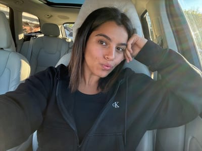 A female individual is seated in the driver's position of a vehicle in Lawrence Township, United States. She has dark hair and a light complexion, wearing a black full-zip hoodie with a white "cK" logo on the left chest, over a black undershirt. Her right hand is positioned against her right cheek, and her lips are pursed as she looks forward. The vehicle's interior features light-colored leather seating and a panoramic sunroof. Bright natural light enters the cabin from the passenger side, causing lens flares and illuminating blurred exterior elements, consistent with a daytime, residential setting.