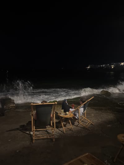 The image depicts a nighttime scene at a waterfront establishment in Naousa, Greece. Two individuals are seated in canvas deck chairs, facing the sea. The person on the right is holding a drink and looking towards the water. Waves are crashing against a rocky shoreline in the foreground, creating a dynamic splash of white water. In the background, lights from a coastal town are visible, suggesting a populated area. Small tables are placed near the chairs, with drinks and what appears to be an ashtray on one of them. The overall atmosphere is relaxed, with the sound and sight of the sea dominating the environment.