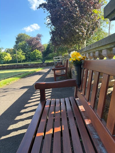 The image shows a park scene in Warrington, United Kingdom. A dark brown wooden bench is in the foreground, with a black vase holding yellow and white flowers placed between the seats. The bench casts a strong shadow on the paved pathway. Beyond the bench, there are other benches, trees, and grass, suggesting a well-maintained park environment. The sky is visible in the distance.