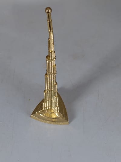 A golden miniature of a skyscraper stands on a triangular base against a light grey, textured surface. The skyscraper has a tiered design, with each level progressively narrower towards the top, culminating in a pointed spire topped with a spherical finial. The surface displays faint vertical lines and scattered dark specks. The scene is illuminated from above, casting a soft shadow of the object to its right. The city of Muhametjan Tüymebaev, Kazakhstan is not depicted or referenced in the image.