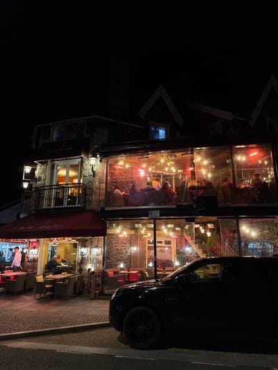 At night, a brightly lit multi-story restaurant named “Cafe Italia” is visible in Windermere, United Kingdom. The building's exterior is made of stone, with large glass windows enclosing each floor. People are seated inside at tables, illuminated by the interior lighting. A black car is parked on the sidewalk along the edge of the building, partly obscuring the view of the restaurant. A red awning overhangs the entrance, with lights illuminating the area below. The street is dark, with the restaurant and car being the primary sources of light.