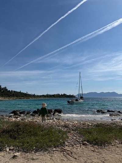 This image depicts a coastal scene on the island of Aegina, Greece, likely a beach or cove looking westward across the Saronic Gulf. In the foreground, a small child with light-colored, medium-length hair, seen from behind, stands on a rocky and pebbly shoreline. The child wears a dark green long-sleeved top and light-colored striped pants, facing towards the water. The immediate ground is a mix of dry soil, sparse green and reddish low-lying vegetation, and scattered dark rocks and pebbles leading to the turquoise-blue sea.

In the midground, a white monohull sailboat with a single mast is positioned in the clear, rippling turquoise water. Two individuals are visible on the boat; one figure in a white shirt stands near the mast, and another is partially visible further aft. A small outboard motor is mounted at the stern.

The background features landmasses under a clear, bright blue sky traversed by several prominent white aircraft contrails and some faint wispy clouds. To the left, a green, tree-covered hillside with scattered residential buildings rises. To the right, across the water, a more distant landmass with rugged mountains (likely the Peloponnese or Methana peninsula) is visible. A distinct, slender, dark-colored monument or pillar stands atop a headland in the right-middle distance. Sunlight glints off the water in the middle ground, indicating a bright day.