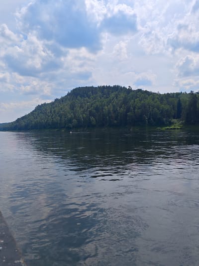 The image shows a wide, dark river flowing in front of a densely forested hill. The sky above is partly cloudy with dramatic white clouds against a blue backdrop. On the far bank, the hill is covered with a thick canopy of green trees, with the top of the hill sloping downwards towards the right. The water surface is textured with ripples, reflecting the sky and the surrounding greenery. In the middle distance on the river, a small, white object, possibly a buoy or a small boat, is visible. The foreground shows the edge of a dark surface, likely a dock or the side of a boat, which is out of focus. The scene appears to be a natural landscape.