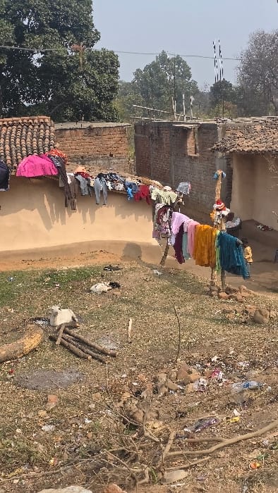 A rural scene depicts a cluster of modest dwellings with terracotta tiled roofs. Clotheslines are strung between poles, laden with brightly colored garments. A dirt yard is strewn with debris, including plastic bottles and scattered trash. Cut firewood is piled near the edge of the yard. Trees with bare branches frame the background, suggesting a season of transition.