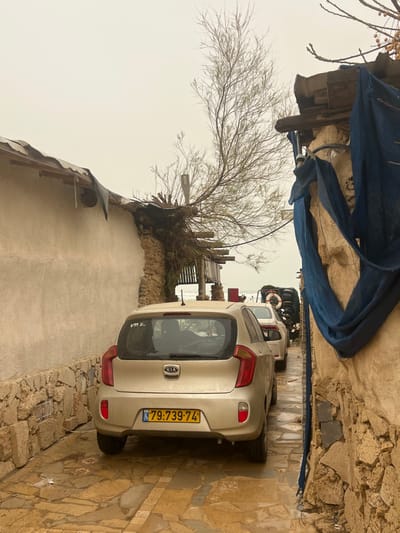 The image depicts a narrow, wet alleyway in Jisr az-Zarqa, Israel, leading towards the sea under an overcast sky. In the foreground, a light gold Kia hatchback, license plate number 79:739-74, is parked facing away from the camera. A white sedan is parked behind it, followed by another vehicle and stacked objects, possibly tires, with a white lifebuoy visible on the right.
The left side of the alley features a wall with a stone base and a stucco upper section, topped by a corrugated metal roof. A bare, deciduous tree with thin branches grows upwards from behind this structure, next to a streetlamp. The right side of the alley is defined by a rough stone wall with an overhang of wooden beams, from which sections of blue fabric are draped. The ground is paved with irregular flagstones, appearing wet in various areas. The sea, characterized by grey, choppy water, is visible at the end of the alley. No individuals are present in the scene.