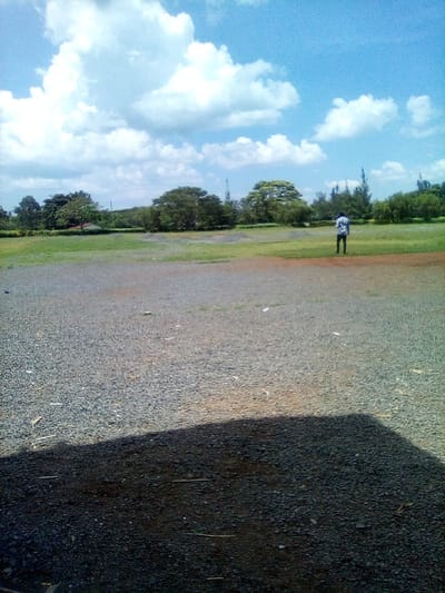 This outdoor image captures a broad landscape under a bright, partly cloudy sky. The foreground is dominated by a large, unpaved area covered in dark grey and brown gravel or small stones, with a distinct dark shadow cast across the bottom left, suggesting an unseen object or structure just out of frame. Beyond the gravel, a wide expanse of green grass stretches into the mid-ground.
A solitary male figure stands in the mid-ground, facing away from the camera, wearing a light-colored, possibly patterned, shirt and dark trousers. He appears to be looking towards the horizon. The background features a line of lush green trees of various sizes and species, forming a natural boundary against the sky. The sky itself is clear blue with numerous fluffy white cumulus clouds scattered across it. The strong sunlight and clear shadows indicate it is daytime. The setting suggests an open, natural area, possibly a park, large field, or cleared ground, consistent with a rural or semi-rural environment in West Sakwa ward, Kenya. There is no visible text in the image.