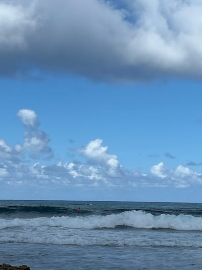 This is a daytime photograph capturing a coastal scene in Le Moule, France. The upper portion of the frame features a bright blue sky partially covered by large, diffuse grey clouds dominating the top section, with scattered white cumulus clouds visible below them. The lower half depicts the ocean, characterized by dark blue-green water transitioning to lighter tones in the foreground. A significant white-capped breaking wave is prominent in the mid-ground, with smaller waves and ripples closer to the viewer. A dark, irregular formation, possibly rocks or coral, is partially visible in the bottom-left corner of the foreground. Two individuals are present in the ocean: one, appearing as a dark head, is partially submerged further out to the left of center, beyond the main breaking wave. A second individual, wearing what appears to be a reddish garment, is also partially submerged, positioned to the right of center and slightly closer than the first. Both individuals are static within the water.