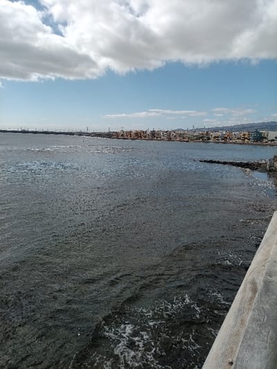 This image presents a wide coastal view of Arinaga, a locality within the municipality of Agüimes, Gran Canaria, Spain. The scene is captured from an elevated perspective, evidenced by a light-colored, textured concrete or wooden railing partially visible along the lower right diagonal edge of the frame.

The upper portion of the image displays a sky with large, irregularly shaped white cumulus clouds across a bright blue expanse. The majority of the frame is occupied by the dark blue-grey waters of the ocean, which appear choppy with numerous small ripples and localized patches of white foam, particularly near the foreground and where the water meets the land on the right.

Along the distant horizon, a developed coastline stretches across the middle ground from left to right. This shoreline features a dense collection of buildings, varying in height and exhibiting exteriors in pastel shades such as tan, white, light blue, and green. A low, dark rock barrier or breakwater extends from the right side of the shoreline into the water. Behind and interspersed with these structures, a prominent array of numerous wind turbines is visible, extending along the coast and into the far distance on the left, suggesting an active wind farm. Further to the extreme left in the distance, larger industrial structures, potentially port cranes, are discernible against the horizon. No human figures are present in the image.