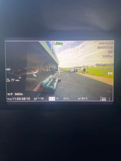 The image displays a camera monitor showing an outdoor scene at the Chonas-l'Amballan airfield in France. In the immediate foreground, a three-seater teal-colored leather sofa is positioned on dark tarmac, with a small black object resembling a case or box situated to its right. Red lines are painted on the tarmac surface, indicating designated areas or pathways.
In the midground, a large aircraft hangar with a green roof and light blue lettering is visible on the left. Inside the hangar, at least two small propeller aircraft are parked, one with a red and white livery and another primarily white. Several individuals are clustered near the hangar entrance. Further to the right, on the tarmac and adjacent grassy area, a larger white propeller aircraft with a blue stripe is parked. A small group of people are gathered around this aircraft, appearing to observe or interact with it.
The background features a partially cloudy sky and green landscape beyond the airfield. The scene is observed through a camera's digital display, evidenced by technical overlay information including "Stby," "DC IN 19.8 V," "3840x2160" resolution, "25P" frame rate, "XAVC-I" codec, "SG3C/SLOG3" picture profile, "LUT s709," "Base 800," "LA 77 min," "W:P 5600K," "TCG 11:00:38:10," "ND 1/19," "F2.8 H:", "800EI," and "4180.0," along with a battery indicator and a central crosshair.