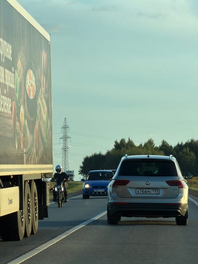 The image captures a highway scene on a clear, possibly late afternoon or early evening day, with a pale blue sky and a few wispy clouds. The foreground is dominated by the side of a large, light-colored truck with a colorful advertisement on its trailer, depicting food items like vegetables and what appears to be a whole cooked chicken or similar round food. The truck's wheels are prominently visible on the left side of the frame.

In the middle ground, a motorcycle with a rider wearing a white helmet and dark clothing is traveling in the same direction as the truck. The motorcycle's headlights are illuminated. To the right of the motorcycle, a blue hatchback car is seen, also in motion. The license plate of the blue car appears to be K882YY 716.

Further ahead, a silver Volkswagen SUV is driving in the right lane. Its rear is facing the camera, and its license plate reads A766MC 799 RUSSIA. The SUV has roof rails.

The background consists of a dense line of green trees and a tall electricity pylon with power lines stretching across the sky. The road itself is asphalt, marked with a solid white line on the left and a dashed white line to the right, indicating multiple lanes. The environment appears to be rural or on the outskirts of a populated area, given the presence of the highway, trees, and pylon. The image is captured from a vehicle traveling on the same highway. The location is identified as Togashevo, Russia.