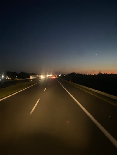 The image depicts a twilight scene on a highway in the vicinity of Węgrzce Wielkie, Poland. The sky is a gradient of deep blue to orange near the horizon. A thin crescent moon is visible in the upper right portion of the sky. The highway stretches into the distance, with a central lane marker and white lines delineating the edges of the lanes. Several vehicles, appearing as bright points of light, are traveling in the distance. Traffic cones, some with illuminated orange tops, are present on the side of the road. Silhouetted trees and power lines line the right side of the highway, with some distant lights suggesting buildings or infrastructure. The overall atmosphere is that of a highway journey at dusk or dawn.