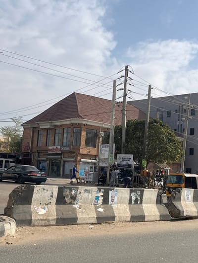 A two-story brick building with a brown roof stands beside a busy street. Overhead power lines intersect the sky, connected to concrete poles. A sign reading "Al-Assaar Academy" is visible amidst other commercial signs. People are gathered on the sidewalk near the storefronts. A black car is parked at the curb. A yellow construction vehicle sits in the background. The scene is bathed in sunlight under a partly cloudy sky.