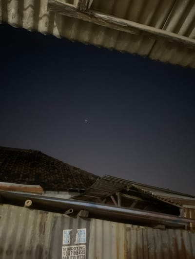 The media file captures an outdoor scene at night, likely in a residential or industrial area of Anggaswangi, Indonesia. The image is dominated by a dark, twilight sky with a subtle gradient, suggesting it is either dusk or dawn, or late evening under minimal light pollution.
Two structures flank the sky, both appearing to be made of corrugated metal and possibly wood. On the left, a weathered, ribbed surface runs diagonally across the frame, creating a sense of depth and framing the sky. On the right, another corrugated structure is visible, with a more textured, possibly brick-like surface beneath the metal sheeting. This right-hand structure has some visible writing on it.
A single, bright point of light is visible in the sky, which could be a star or a planet. It is the only discernible celestial body in the frame.
The overall mood is quiet and still, with no signs of human activity. The weathered appearance of the structures suggests a rustic or possibly aging environment. The lack of distinct atmospheric conditions implies clear or partly cloudy weather.
Visible text on the right-hand structure includes:
- "SEDOT WC"
- A phone number: "085 71666 4545"
- Another partial phone number: "0854 030 11520"
- Other partial labels and numbers, like "085 74737", "08 6339". These are likely related to local services or identification.