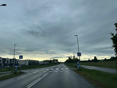 The image depicts a road in Tiskre, Estonia, under a cloudy sky. The road is paved with asphalt and has white lane markings. A pedestrian crossing is marked with white diagonal stripes. On the left side of the road, there are modern, multi-story buildings with dark facades and large windows. There are also streetlights and street signs present. On the right side of the road, there is a grassy field with some trees and vegetation. The overall scene suggests a suburban or semi-rural setting.