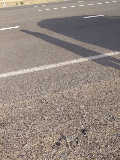 A low-angle, eye-level shot shows an asphalt road in Қапанбұлақ ауылдық округі, Kazakhstan. White dashed lines mark the lanes of the road, with a solid white line closest to the camera. The road is bordered on the left by a dirt shoulder covered in small stones and debris, and on the right by more asphalt. The shadow of a vehicle, likely a car or bus, stretches across the road from left to right, indicating a sunny day. The background shows a hint of dry vegetation and a distant landscape.