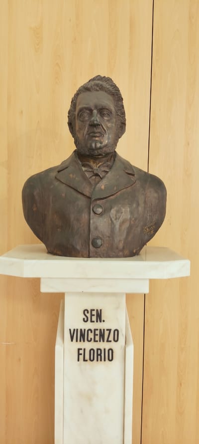 A dark bronze bust of a man is displayed on a white marble pedestal against a light wood-paneled background. The bust depicts a man with neatly combed hair, a beard, a collared jacket with two buttons, and a bowtie. There is a signature and a date, "ARMONIO 1986", inscribed on the right side of the bust. The marble pedestal has the name "SEN. VINCENZO FLORIO" inscribed in black lettering on its front. The bust is positioned centrally on a white marble shelf, which rests on the pedestal. The wood paneling behind the bust shows vertical grain patterns and appears to be made of light-colored wood. The lighting is even, suggesting an indoor setting. This bust is located in the Museo dell'Insalata or the Florio Museum, in Marsala, Italy.