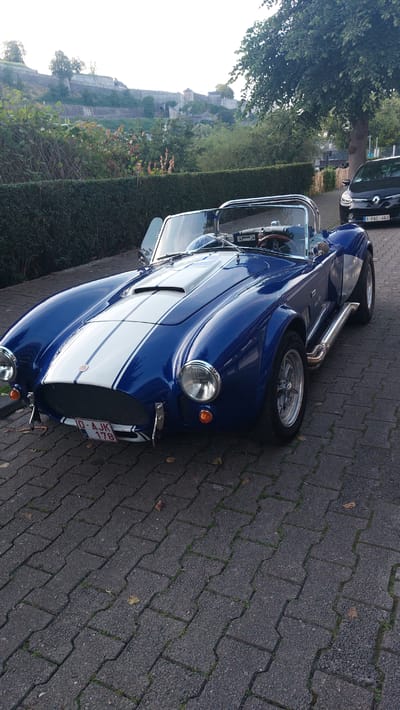 In the city of Namur, Belgium, on a paved surface, a blue convertible sports car with white racing stripes is parked. The car has its driver's side door open. In the background, a black car is parked, and further in the distance, a historical fortress or castle structure is visible on a hillside, surrounded by trees and greenery. The scene is set outdoors, likely on a street or driveway.