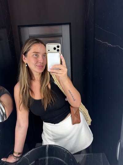 The image shows a young woman with long, blonde-brown hair, wearing a black tank top and a white skirt. She is holding a smartphone and taking a selfie in a bathroom with dark marble walls. She has a gold necklace and earrings, a tattoo on her arm, and a woven bag with a brown glasses case attached. The location is identified as Укромновское сельское поселение, Ukraine, though there are no visual cues in the image to support this specific location. The woman is looking at the camera with a slight smile.