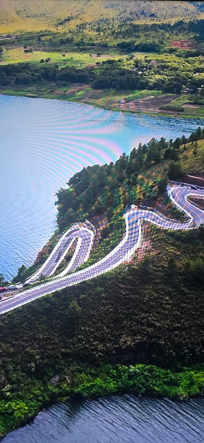 The image is an aerial view of a winding road snaking up a hillside next to a body of water. The road has multiple hairpin turns and is lined with white barriers. There are a few vehicles visible on the road. The hillside is covered with trees and vegetation, and in the distance, there are rolling hills and fields. The water appears to be a lake or reservoir. There is no indication of the city of Lippstadt, Germany, in this image.