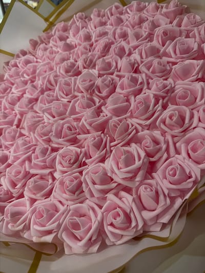The image displays a close-up, angled view of a dense arrangement of numerous light pink artificial roses. The roses, likely made of foam or fabric, are uniformly shaped with tightly spiraled centers, creating a textured surface. They are packed closely together, filling the majority of the frame.
The roses appear to be part of a larger decorative item, possibly a large bouquet, a heart-shaped display, or a gift arrangement. Along the right and bottom edges of the main image, there are hints of a lighter, possibly off-white or cream-colored, wrapping or backing material, accented with thin, wavy golden lines or ribbons. This suggests the roses are presented within a decorative context.
There is no activity taking place, as the image captures a still life. The soft pink color of the roses typically symbolizes gentleness, admiration, joy, and love, suggesting the item is a gift or decoration for a special occasion such as a birthday, anniversary, or Valentine's Day.
The lighting is soft and even, highlighting the contours of the individual rose petals and the slight shadows cast by each flower, adding depth to the composition. The time of day and weather are indeterminate, suggesting an indoor setting. There are no specific visual cues within the image itself to indicate the location as Cherni Vrah, Bulgaria. No visible text is present in the image.