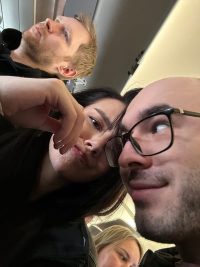 The image depicts an interior scene with four individuals, captured from a low angle, suggesting a selfie or close-up perspective. The specific location in the city of Stolbishche, Russia, cannot be determined from the visual evidence; the environment appears to be an interior space, possibly a cabin of a vehicle like an airplane or train, indicated by a low ceiling and overhead paneling.

In the foreground right, a light-skinned male with a bald head and short stubble beard is partially visible in profile, facing left. He wears black-framed glasses with gold-colored accents. His right eye is visible, looking upward and slightly to the right. His lips are slightly parted.

Positioned behind and to the left of the foreground male, a light-skinned female with long dark hair is prominently featured, looking directly towards the camera with a slight pout or wry expression. Her right hand is raised with fingers lightly curled, appearing to be near her chin or mouth. She exhibits clear nail polish on her fingernails and is wearing dark clothing.

In the upper left background, a light-skinned male with short blonde or light brown hair and a beard is visible in profile, facing left and looking upward. He wears a dark top, possibly a hoodie.

Partially visible in the lower right, behind the foreground male, is a light-skinned person with blonde or light brown hair, looking upward and to the right. Only the forehead, right eye, and a portion of the cheek are discernible.

The overall environment suggests an artificial light source from above, illuminating the subjects within a confined, functional space. There are no explicit interactions between the individuals, though their close proximity implies a shared space or group.
