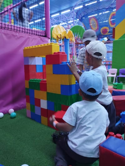 In an indoor play area in Talas City, Kyrgyzstan, three children are engaged with a large structure built from oversized, colorful building blocks. The structure is composed of red, yellow, blue, green, and white blocks. In the foreground, a child wearing a blue cap and white t-shirt is kneeling and appears to be interacting with a small toy car. Behind this child, another child, wearing a white cap and t-shirt, is standing and reaching towards the block structure. Further back, a third child, partially obscured, is also near the blocks. The play area has a green carpeted floor with scattered colorful balls and soft play modules. In the background, vibrant netting and play equipment are visible, suggesting a dedicated children's entertainment facility.