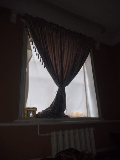 The image shows a dimly lit interior, possibly in Kuvasay, Uzbekistan. A window with a draped, tasseled curtain is the focal point. A partially obscured white curtain hangs behind the draped one. A yellow plastic container sits on the windowsill to the left. Below the window, a radiator is visible. The walls are a muted brown color. The overall lighting is poor, making details difficult to discern.