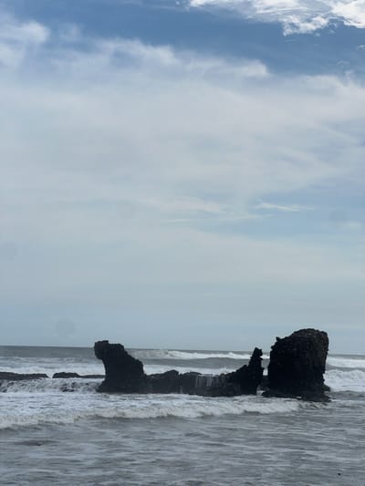 A rocky formation protrudes from the turbulent, grey ocean waters at El Tunco beach, El Salvador. The rocks are dark and jagged, with waves crashing around them, creating white foam that contrasts with the darker water. The sky above is overcast with a mix of grey clouds and patches of lighter sky. The scene is devoid of human presence.