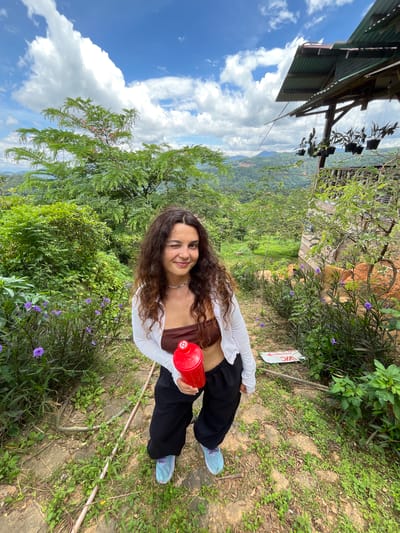 A woman with long, curly brown hair is standing outdoors, winking at the camera and holding a red water bottle. She is wearing a brown strapless top, a white cardigan, black pants, and light blue sneakers. The background shows a lush, green landscape with rolling hills and mountains, indicative of the Sơn Điền area in Vietnam. A rustic, open-air structure with a corrugated metal roof and hanging plants is visible to the right. The ground is covered with dirt, gravel, and patches of green vegetation, including small purple flowers. The sky is blue with scattered white clouds.