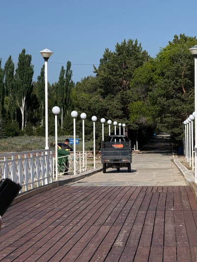 In Koshkol', Kyrgyzstan, a scene unfolds on a paved pathway lined with white railings and spherical lamps. A man, wearing a dark vest over a blue shirt and yellow gloves, is engaged in emptying a large green garbage bin into a dark-colored, three-wheeled vehicle. The vehicle is parked on the pathway, facing away from the viewer. The pathway itself is surrounded by lush green trees and a clear blue sky. The foreground is dominated by a wooden deck. In the background, a blue sign with white text and an icon is visible.