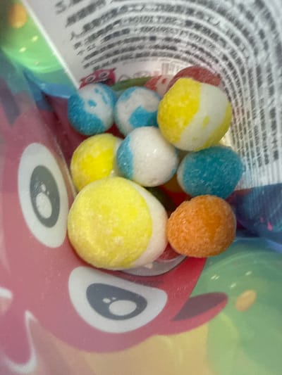 The image displays a close-up view of several brightly colored, round candies scattered inside a pink bag with a cartoon character print. The candies are a mix of white, yellow, blue, and orange, some with multiple colors. The bag appears to be made of a plastic material and has a glossy finish. In the background, there is a blurred view of text on a white surface and a patterned interior of the bag. The scene does not contain any information or context related to Yakovlevskoye, Russia.