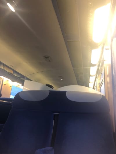 The image depicts the interior of a train carriage, in the context of Hochfelden, France. The viewpoint is from a low, seated position, looking forward and upward within the carriage. In the foreground, the upper backs of two dark blue upholstered train seats are partially visible. Directly beyond, a row of seats features dark blue upholstery with two distinct lighter gray oval-shaped inserts on each backrest. These seats are separated from the next section by a clear, curved acrylic or glass partition. Above this partition, the crown of a person's head with dark hair is discernible. The ceiling is light-colored, featuring multiple recessed circular lights and two visible smoke detectors. Along the right side of the carriage, a series of bright, warm-toned rectangular overhead light fixtures illuminate the space, with one particularly intense light source causing significant lens flare and overexposure in the upper right. A partial view of another dark blue seat is visible on the far left.