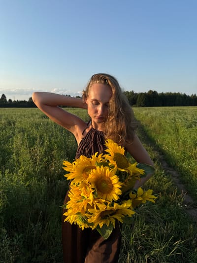 A young woman with long blonde hair is pictured outdoors, holding a bouquet of sunflowers. She is wearing a dark brown dress with a ruched neckline. Her eyes are closed, and her head is tilted back slightly, with her left arm raised behind her head, suggesting a relaxed or pensive mood. The sunflowers are bright yellow and fill the right side of the frame, obscuring part of her body.
The setting is a grassy field under a clear blue sky. In the background, a line of trees is visible along the horizon, and a dirt path can be seen to the left of the woman. The lighting suggests it is late afternoon or early evening, with the sun casting a warm glow on the scene. The overall atmosphere is serene and natural. The image appears to have been taken in Aksaitovo, Russia. There is no visible text in the media file.