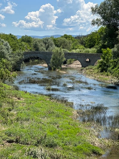 An eye-level, medium shot captures a tranquil scene of a stone bridge with two arches spanning a shallow, flowing river under a partly cloudy blue sky. Lush green foliage lines both banks of the river, with trees forming a dense backdrop behind the bridge. The water reflects the sky and the surrounding greenery, with some reeds and vegetation growing within the riverbed. In the foreground, the grassy bank slopes down towards the water. The image is devoid of any human presence or activity. The stone bridge appears ancient and weathered, suggesting historical significance, and is likely located in the vicinity of Debelets, Bulgaria, given the description of the location.