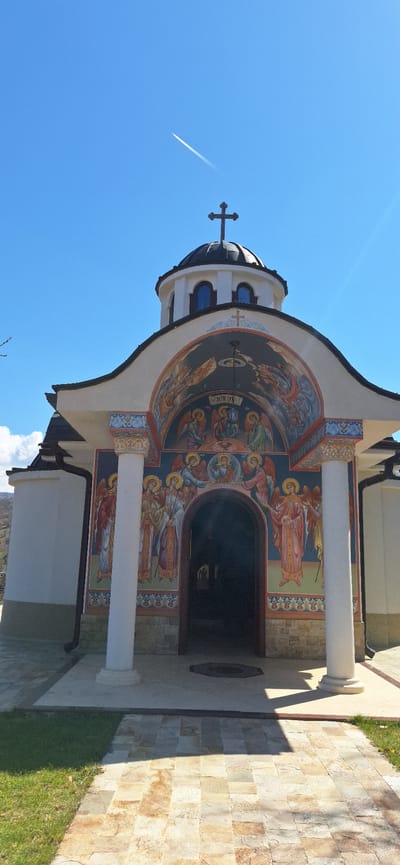 Here's a description of the image:
The image captures a section of what appears to be an Orthodox church. The building is decorated with religious iconography, depicting what appear to be angelic figures and other religious motifs painted around the entrance and under the domed roof. The entrance is framed by white columns, and there's an arched doorway leading inside. To the right of the building, there's a paved walkway or patio made of stone. The sky is clear blue with a contrail from a plane visible. The scene depicts the church located in the city of Elenovo, Bulgaria.