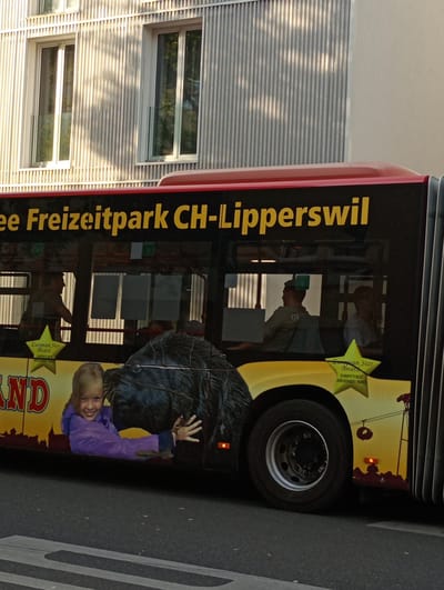 The image shows the side of a bus with advertisements. The bus has a yellow and black color scheme with yellow lettering that reads "ee Freizeitpark CH-Lipperswil". There is a graphic of a girl in a purple jacket hugging a black furry creature, possibly a bear or a beaver, on the side of the bus. A yellow star with "European Star Award" and "EUROPE'S BEST AMUSEMENT PARKS" is also visible. Several windows on the bus reveal silhouettes of people inside, including one person appearing to be a driver. The bus is parked on a street in front of a modern building with vertical siding and windows. The street has a pedestrian crossing in the foreground. The overall scene suggests the bus is part of a transportation service for a theme park, possibly located in or near Constance, Germany.