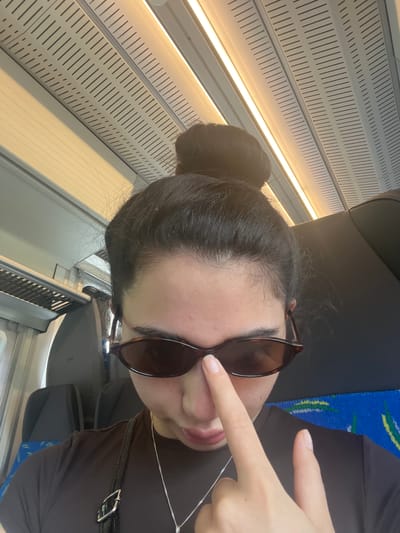In the provided image, a young woman is captured from the chest up, her face partially obscured by sunglasses and a finger resting on her nose. She is wearing a dark brown t-shirt and a silver necklace. Her hair is styled in a messy bun. She appears to be inside a train, indicated by the rows of seats in the background, one with a blue and yellow pattern. The ceiling of the train carriage features illuminated panels and ventilation grilles. The context suggests she is traveling within the city of Vejprnice, Czechia.