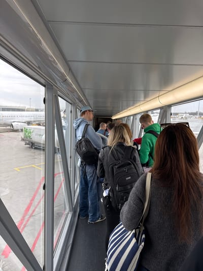 The photograph was taken in Oberding, Germany, inside an airport jet bridge. The image shows a line of passengers boarding or disembarking an airplane, likely a Gate Gourmet plane based on what can be seen outside the window. Passengers are wearing a variety of clothing. The windows of the jet bridge are visible, providing a view of the tarmac. The jet bridge is lined with gray panels. The image is taken from the perspective of someone standing in the line of passengers.