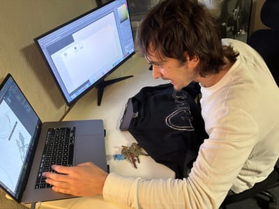 The scene depicts an indoor workspace located in the city of Oberhausen, Germany. A single adult male is shown from the upper torso, seated at a light-colored desk, facing left and slightly downwards. He has medium-long, dark brown hair with a longer length at the nape and sides, and a light goatee. He wears a plain white long-sleeved t-shirt; his mouth is slightly open. His right hand rests on the keyboard of an open silver Apple MacBook Pro laptop, whose screen displays what appears to be a topographical map or design software with landmass outlines and contour lines. To the right of the laptop, a black Acer monitor shows a graphical user interface resembling design software like Adobe Photoshop, featuring toolbars and an empty white canvas or file browser. A dark t-shirt or sweatshirt with a faded white graphic is folded and placed on the desk between the laptop and the male. A set of metal keys with a small light blue plastic attachment lies on the desk to the right of the clothing item. A dark office chair with a black mesh back is partially visible behind the male.