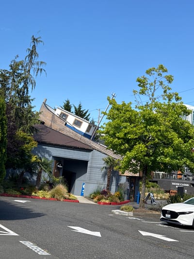The image shows the exterior of a building that has a boat placed on its roof, giving the illusion that the boat is sinking into the structure. The boat is mostly wood-colored with a white and blue cabin. The building itself is gray with a brown, corrugated metal roof. The scene is set on a sunny day with a clear blue sky.
The building is identified by a sign that reads "Dockside." In the background, another building has signage for "SILVER CLOUD HOTEL." The location is in Seattle, Washington, USA.
In the foreground, there is an asphalt road with white directional arrows painted on it. A white car is parked on the right side of the road. The landscaping around the building includes trees, palm trees, and various plants. The overall impression is a unique architectural design or a themed establishment.