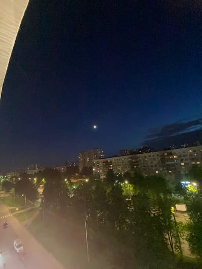 The image depicts a nighttime view from an elevated position in Borisova Griva Massiv, Russia. The scene is divided between the night sky and a cityscape. In the sky, a small, bright celestial object, likely the moon or a bright star, is visible. The cityscape consists of tall buildings with illuminated windows and green areas possibly representing parks or trees. Streetlights are scattered throughout the scene. The foreground features a light-colored structure, possibly a building ledge or balcony. A road with cars can be seen in the distance.
