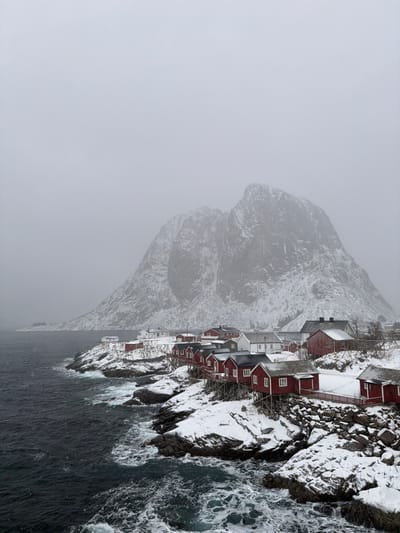 The image depicts a winter scene in Hamnøya, Norway, under an overcast grey sky. A cluster of traditional red and white wooden houses, many built on stilts, line a snow-covered rocky coastline on the right and center-right. The dark, choppy ocean with white breaking waves occupies the left foreground, contrasting with the snow. Wooden structures, likely drying racks, are visible between the buildings. A massive, steep, snow-dusted mountain dominates the background, its peak partially obscured by the low clouds. No individuals are visible in the scene. The ground, rooftops, and mountain slopes are covered in snow, indicating cold weather conditions.