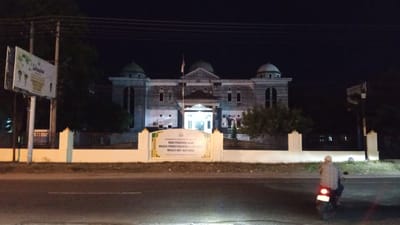 This is a nighttime, eye-level, medium shot of a building and its surroundings in Cotbadatunong, Indonesia. The building is large and has a classical architectural style, with domes and a central portico. It is illuminated by lights, making its facade visible against the dark sky. In front of the building is a white wall with decorative pillars and a prominent sign. The sign, in Indonesian, appears to be from the local government, mentioning the Education Department and cultural/religious councils. To the left of the building, a billboard stands tall, displaying text and images, also lit up.
In the foreground, a paved road runs across the image. On the road, a person on a motorcycle is visible from behind, heading away from the camera. The person is wearing a checkered shirt and a helmet. The overall scene suggests a calm, ordinary evening in a populated area, possibly a town or city center, given the presence of public buildings and infrastructure like roads and billboards. The time of day is clearly night, and the weather appears to be clear, allowing for good visibility of the illuminated structures.