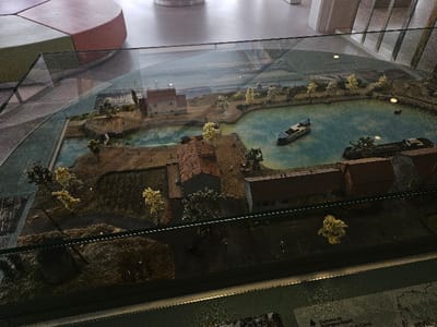 The image displays a detailed miniature diorama, presented under a reflective glass case, depicting a landscape within the city of Avignonet-Lauragais, France. A central, light blue body of water, consistent with a canal basin, dominates the scene. Two miniature vessels are on the water: a multi-decked boat, predominantly white with a blue hull, is positioned centrally, and a darker, flat-decked barge is docked along the right bank.
The surrounding land is textured with brown and green, representing earth and vegetation. Numerous miniature trees with light green and yellow foliage are distributed across the land, particularly along the water's edge and adjacent to structures. Several buildings with reddish-brown tiled roofs and light-toned walls are visible, including a prominent structure on the upper-left bank, another centrally located on the lower bank, and a row of connected buildings extending along the lower-right bank.
Dark pathways or roads traverse the land areas, and a thin, dark line resembling a railway track runs parallel to the row of connected buildings in the lower-central section. Numerous minuscule human figures are scattered across the diorama, suggesting habitation and activity, though individual details are indistinct. The glass display case reflects external elements, including a curved, multi-colored seating object in the upper left, and partially obscures an informational panel in the lower right foreground.