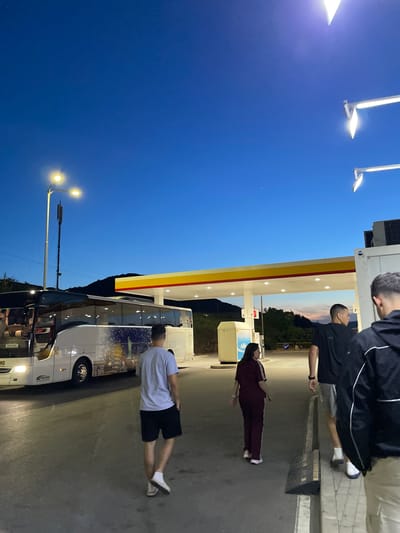 The image depicts a gas station in Kalotina, Bulgaria at dusk. A white bus is parked near the station, and several people are standing nearby. One individual is wearing a maroon tracksuit, another a black tracksuit. The gas station is a Shell station, and the sky is a deep blue color. Streetlights are visible in the background.