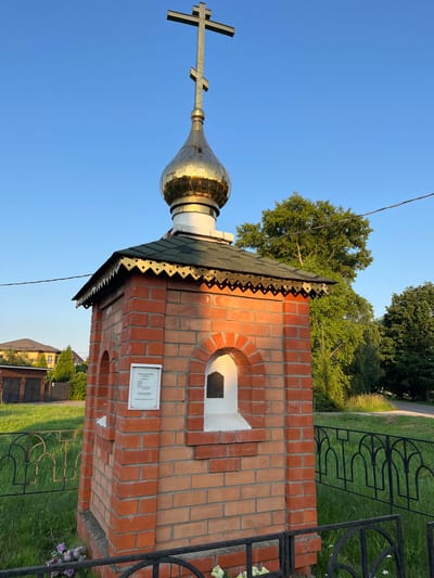 A small, ornate brick chapel stands in a sunlit outdoor setting, likely in Pushkino, Russia. The chapel features a golden dome topped with a cross, and its red brick exterior is adorned with decorative edging under the roof. An arched window with a white sill is visible on the front. A sign with text, though largely unreadable, is affixed to the wall. The chapel is surrounded by a black metal fence, and the ground is covered in green grass, with a path and more trees visible in the background. The sky is a clear, vibrant blue, suggesting it is a sunny day. The overall impression is one of peacefulness and reverence.